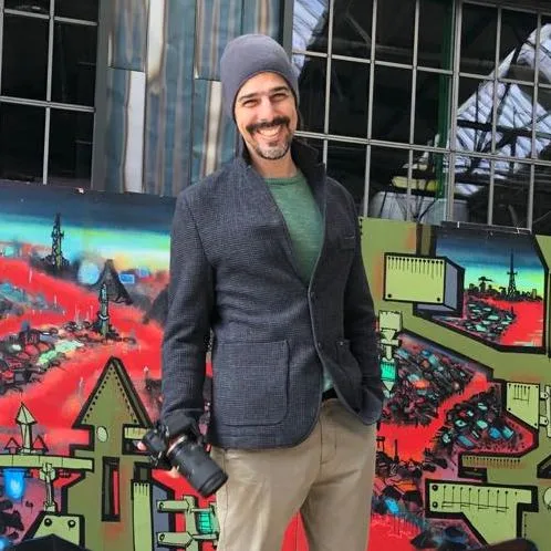Daniel Farjoun sorrindo com uma câmera na mão, em frente a um painel de grafite
